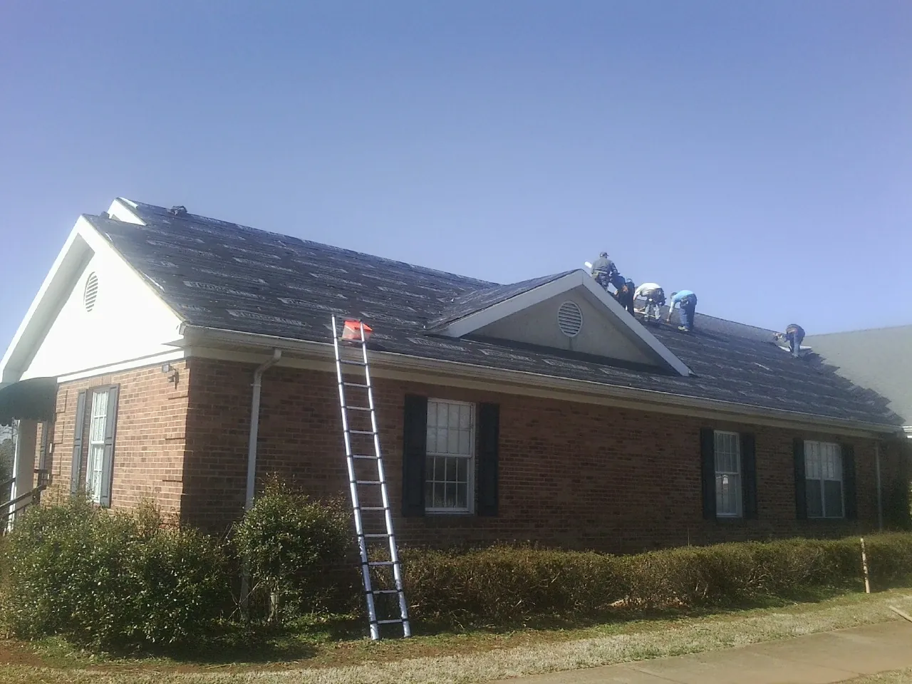 Crew tearing shingles off a brick ranch roof with a ladder set — Emergency Roof Repair work by our crew in Ayer