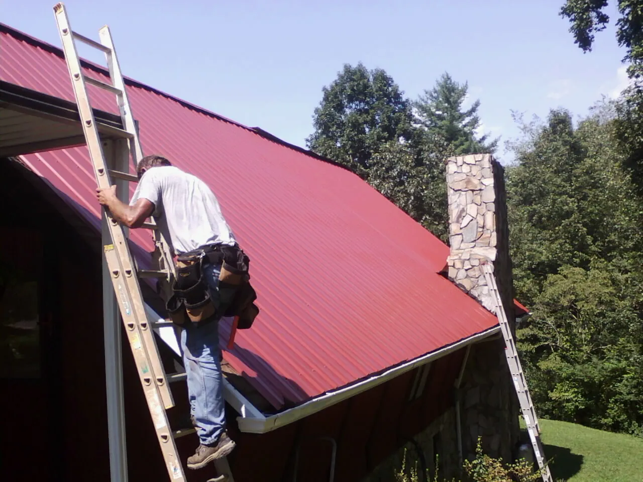 Roofer on a ladder beside a red metal roof and stone chimney — Soffit & Fascia Repair by Roofing Brothers in Ayer