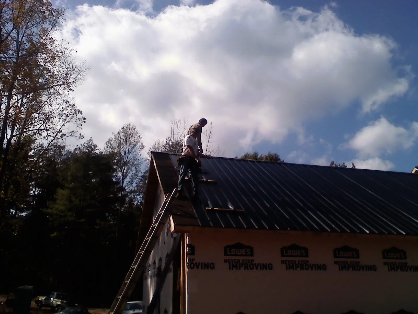 Roofer fastening dark metal panels high against a cloudy sky, from a Roof Inspection job in Ayer